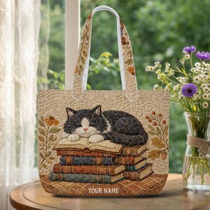 May include: A beige quilted tote bag featuring a black and white cat sleeping on a stack of books. The bag has two handles with a floral pattern and the text YOUR NAME printed at the bottom. The design includes illustrations of flowers.