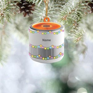 May include: White Christmas ornament shaped like a speaker with a string of colorful lights around the top and bottom. The ornament has an orange light inside and the word "Name" printed on the front.