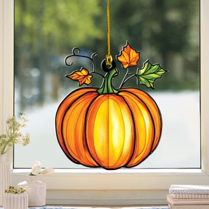May include: A decorative pumpkin window cling with an orange and yellow gradient, black outlines, and green and orange leaves. The pumpkin is suspended by a gold cord, perfect for fall decor. The image is set against a blurred outdoor background.
