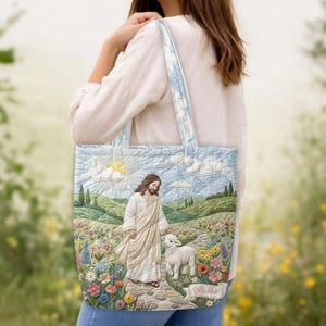 May include: A quilted tote bag featuring a printed scene of a figure in a white robe walking with a lamb on a stone path through a colorful flower field, with rolling green hills, a sun, and clouds in the background and the name Stella printed.