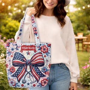 May include: A quilted tote bag featuring a patriotic American flag-themed bow and stethoscope graphic. The design includes red, white, and blue floral patterns and a rectangular label with the text YOUR NAME. The bag has two shoulder straps.