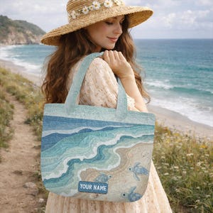 May include: A rectangular fabric tote bag featuring a quilted ocean scene with blue waves, sandy shore, and sea turtles. A rectangular patch at the bottom center displays the text "YOUR NAME" in white letters against a dark blue background.