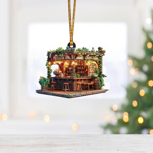 May include: A miniature wooden bar ornament with a sign that reads "The Wishing Well". The bar has a counter with stools, a shelf with bottles, and a green plant hanging from the ceiling. The ornament is decorated with lights and has a gold string for hanging.