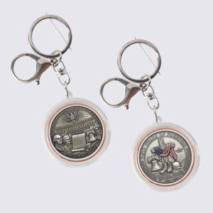 May include: Two keychains with clear plastic cases and silver-tone metal hardware. Each keychain features a round, silver-colored medallion with patriotic imagery and text. One reads "WE THE PEOPLE" and the other reads "250 YEARS OF FREEDOM".