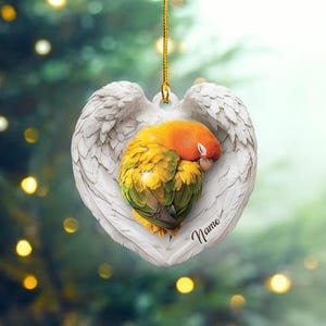May include: A white heart-shaped ornament with angel wings and a sleeping parrot inside. The parrot is yellow, green, and orange. The ornament has the word "Name" written on it.