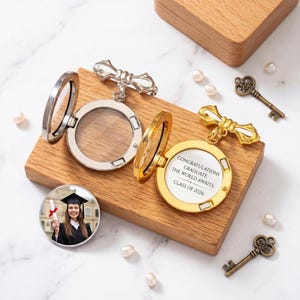 May include: A silver and gold locket with a graduation theme, open to reveal a photo and the message "Congratulations Graduate! The World Awaits. Class of 2026." Decorative keys and pearls are scattered around the wooden display.