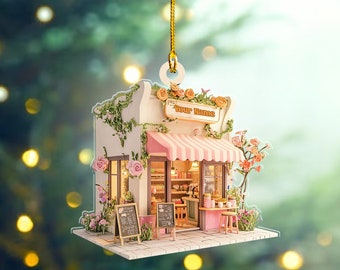 Könnte beinhalten: Ein Miniatur-Puppenhaus-Anhänger in Form eines rosa-weiß gestreiften Ladengeschäfts mit einem Schild, auf dem "Your Name" steht. Die Ladenfront ist mit Blumen und Ranken verziert.