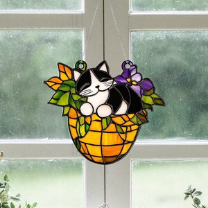 May include: A stained glass suncatcher featuring a black and white cat sleeping in a basket of purple flowers. The basket is orange and yellow with a woven pattern.