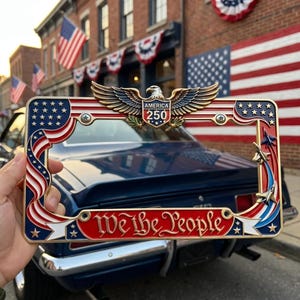 May include: A rectangular metal license plate frame featuring a patriotic design with red, white, and blue colors. The top center displays a golden eagle emblem with the text AMERICA 250. The bottom bar features the text We the People in gold.
