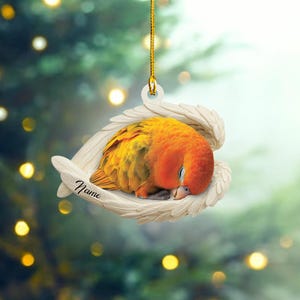 May include: A white ceramic ornament with a sleeping orange and yellow bird inside. The ornament has a pair of white wings and the word "Name" is written on the side.
