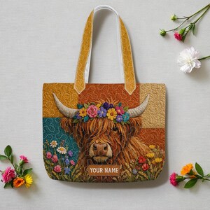 May include: A quilted tote bag featuring a brown Highland cow with a colorful floral crown between its horns. The background is a patchwork of mustard yellow and teal squares. The text "YOUR NAME" is printed in white at the bottom center.