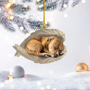 May include: A Christmas ornament featuring a sleeping brown and white dog nestled in a pair of angel wings. The wings are a light beige color, and the dog is curled up, appearing peaceful. The ornament hangs from a gold string.
