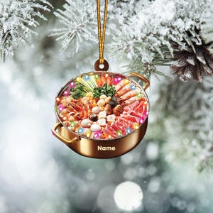 May include: A gold-colored Christmas ornament shaped like a hot pot filled with various ingredients, including shrimp, vegetables, and meat. The ornament is decorated with colorful lights around the rim. The ornament has the word "Name" printed on the bottom.