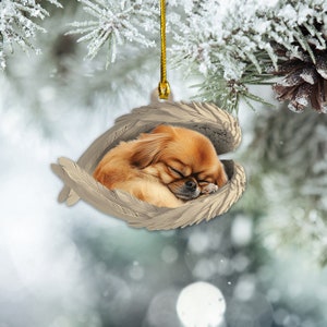 May include: A small, brown dog with closed eyes sleeps peacefully inside a white, sculpted ornament shaped like angel wings. The ornament is a Christmas tree decoration.