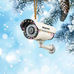 May include: A white security camera ornament with colorful lights strung around it. The camera has a lens and a black base. The ornament has the word "Name" written on it.
