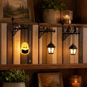May include: Three black metal wall-mounted brackets with decorative scrollwork, each featuring a hanging light fixture. One light is a yellow ghost holding a book, while the other two are black lanterns with silhouettes of witches on the brackets.