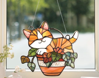 Könnte beinhalten: Ein hängender Bleiglas-Fensteranbau zeigt eine süße orange-weiße Katze, die in einem Blumentopf schläft. Die Katze ist von grünen Blättern und Blumen umgeben.