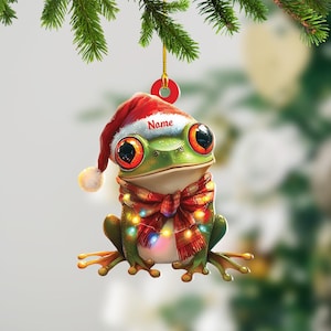 May include: A whimsical Christmas ornament featuring a green frog wearing a Santa hat and a red scarf adorned with string lights. The frog has large, expressive eyes and the word "Name" is printed on the hat. The ornament is hanging from a green branch.
