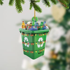 May include: A green recycling bin with a white recycling symbol and a string of colorful Christmas lights wrapped around it. The bin is filled with various bottles and containers. The bin is hanging from a string and has the word "Name" written on the bottom.