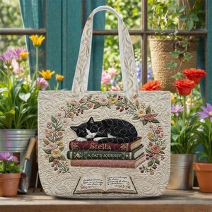 May include: A quilted tote bag featuring a black and white cat sleeping on a stack of three books titled Stella, A Cat's Journey, and Heirloom Dreams, surrounded by an embroidered floral wreath and an open book with printed text at the bottom.