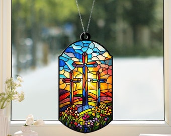 Könnte beinhalten: Ein Buntglasfenster, das mit einem schwarzen Rahmen und einer silbernen Kette aufgehängt ist. Das Bild zeigt drei Kreuze auf einem Hügel mit einem bunten Sonnenuntergang dahinter. Die Kreuze sind rot, gelb und blau. Der Sonnenuntergang ist orange, gelb und blau. Am Fuße des Hügels befinden sich Blumen.