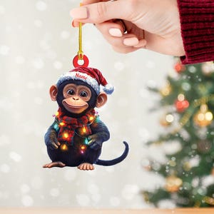 May include: A Christmas ornament featuring a cartoon chimpanzee wearing a Santa hat, a plaid scarf, and string lights. The ornament has a gold string for hanging and the word "Name" on the hat.