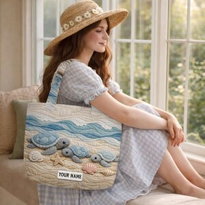 May include: A quilted tote bag featuring a beach scene with three sea turtles, a starfish, and seashells on a sandy shore with blue ocean waves. The bag includes a rectangular white label at the bottom with the black text YOUR NAME.