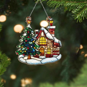 May include: A stained glass Christmas ornament depicting a snow-covered house with a red roof, a lit window, and a small evergreen tree in front. The ornament is hanging from a silver chain.