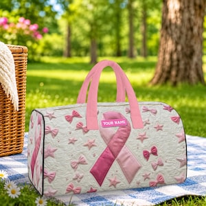 May include: A white duffel bag featuring a pink awareness ribbon, small pink bows, and star patterns. The bag has pink handles and a rectangular pink patch with the text YOUR NAME printed in white. The surface has a quilted texture.
