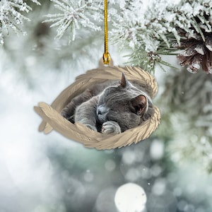 May include: A gray cat ornament sleeping inside a heart-shaped nest of feathers. The ornament is made of ceramic and is perfect for a Christmas tree.