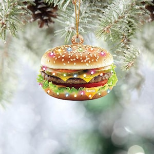 May include: A Christmas ornament shaped like a hamburger with colorful lights around it. The ornament has a sesame seed bun, a beef patty, cheese, lettuce, tomato, and pickles. The ornament says "Name" on the bottom.