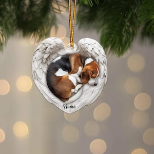 May include: A heart-shaped Christmas ornament featuring a sleeping beagle dog nestled within white angel wings. The dog has brown, black, and white markings. The ornament has a gold string for hanging and the word "Name" is written on the bottom.