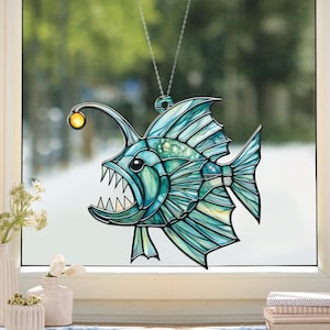 May include: A stained glass-style anglerfish suncatcher. The fish is primarily teal and blue with a yellow lure. It has sharp teeth and is suspended by a silver chain. The suncatcher is displayed in front of a window.