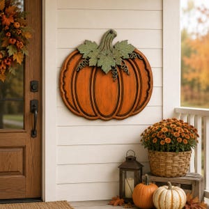 May include: A decorative wall-mounted pumpkin sign featuring a vibrant orange body with dark brown accents, topped with sage green leaves and curled vines. The pumpkin has a textured, ribbed surface and is displayed on a white horizontal paneled wall.