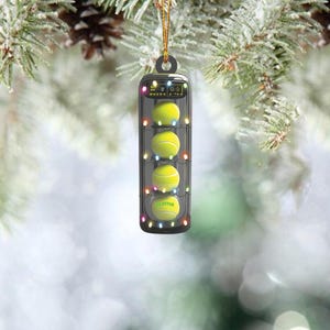 May include: A black and silver Christmas ornament shaped like a tennis ball holder with four tennis balls inside. The ornament has a string for hanging and the text "name" on one of the tennis balls.