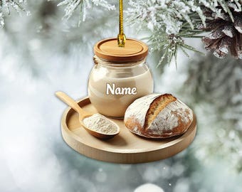 Könnte beinhalten: Ein Holz-Anhänger in Form eines Glases mit Deckel, gefüllt mit einer weißen Substanz. Das Glas trägt die Aufschrift "Name". Der Anhänger wird von einem Holzlöffel mit weißem Pulver und einem runden Brotlaib begleitet. Der Anhänger hängt an einer goldenen Schnur vor einem verschneiten Hintergrund.