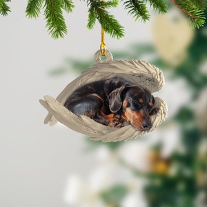 May include: A brown and black dachshund dog sleeping inside a white ceramic ornament with angel wings. The ornament is hanging from a gold string.