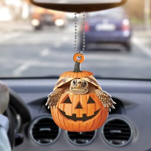 May include: A small turtle is peeking out of a carved orange pumpkin with a smiling face. The pumpkin is hanging from a silver chain, perfect for a car rearview mirror decoration.