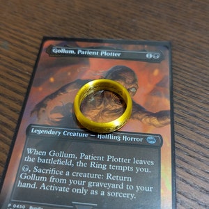 May include: A gold-colored ring with a textured surface, resting on a Magic: The Gathering card featuring Gollum, Patient Plotter. The card text reads: "When Gollum, Patient Plotter leaves the battlefield, the Ring tempts you. Sacrifice a creature: Return Gollum from your graveyard to your hand. Activate only as a sorcery."