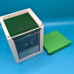 May include: A white deck box with a green deck of cards inside. The top card is a Magic: The Gathering card featuring a blue and green illustration of a creature called "Leinore, Autumn Sovereign". The box is open and a green lid is sitting next to it.