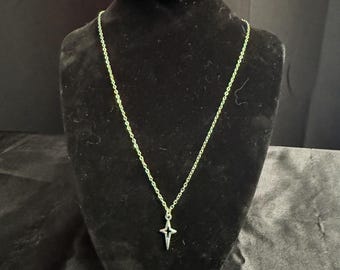 May include: A silver-colored necklace with a star-shaped pendant is displayed on a black velvet bust. The chain is delicate, and the pendant has a four-pointed star design. The background is a dark, solid color.