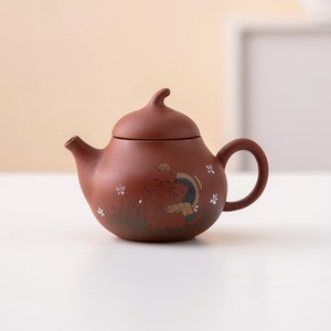 Op de afbeelding: Een bruine theepot met een deksel en een handvat. De theepot is versierd met een geschilderd ontwerp van een meisje dat een kat vasthoudt. Het ontwerp is in tinten bruin, roze en wit.