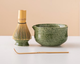 May include: A matcha tea set featuring a green ceramic bowl with a spout, a bamboo whisk, and a bamboo scoop. The whisk is light brown with a green base, and the bowl has a speckled texture. The set is arranged on a white surface.