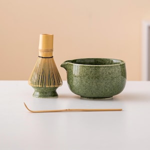 May include: A green ceramic matcha bowl with a bamboo whisk and a bamboo matcha scoop on a white table.
