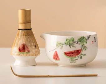 Könnte beinhalten: Ein Matcha-Teeset mit einem Bambusbesen, einer Keramikschale mit Wassermelonen-Illustrationen und einem Bambuslöffel. Der Schneebesen hat einen Bambusgriff und einen weißen Sockel mit Wassermelonen-Design. Die Schale ist weiß mit grünen Blättern und Wassermelonenscheiben.