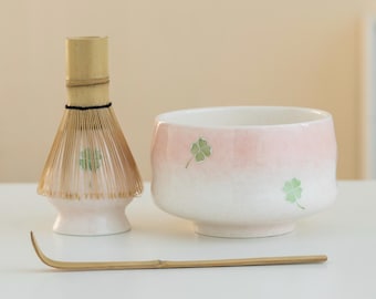 May include: A matcha tea set featuring a pink and white ceramic bowl with green clover designs, a bamboo whisk, and a bamboo scoop. The bowl has a gradient color effect. The whisk is made of bamboo.
