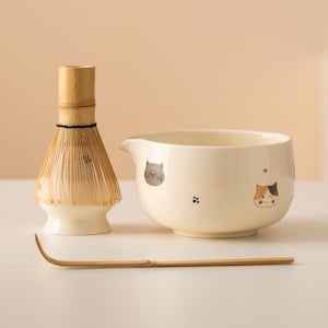 Può includere: Un set da tè matcha con una ciotola in ceramica color crema con illustrazioni di gatti, un frullino in bambù e un cucchiaio in bambù. La ciotola ha un beccuccio. Il frullino e il cucchiaio sono in bambù.
