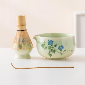 May include: A matcha tea set comprising a pale green ceramic bowl with a spout and blue floral pattern, a bamboo whisk, and a bamboo scoop. Ideal for preparing matcha tea.