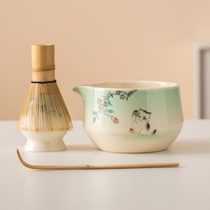 以下が含まれることがあります： 抹茶ティーセット。 竹製の泡立て器、猫のイラストが描かれたセラミックボウル、竹製のスクープが含まれています。 ボウルは淡い緑色のグラデーションと花のアクセントがあります。 泡立て器は天然の竹でできています。