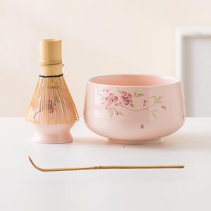 May include: A light pink ceramic matcha bowl and whisk set. The bowl features a floral design in pink and green. The whisk is made of bamboo and has a light pink base. A bamboo scoop is also included.